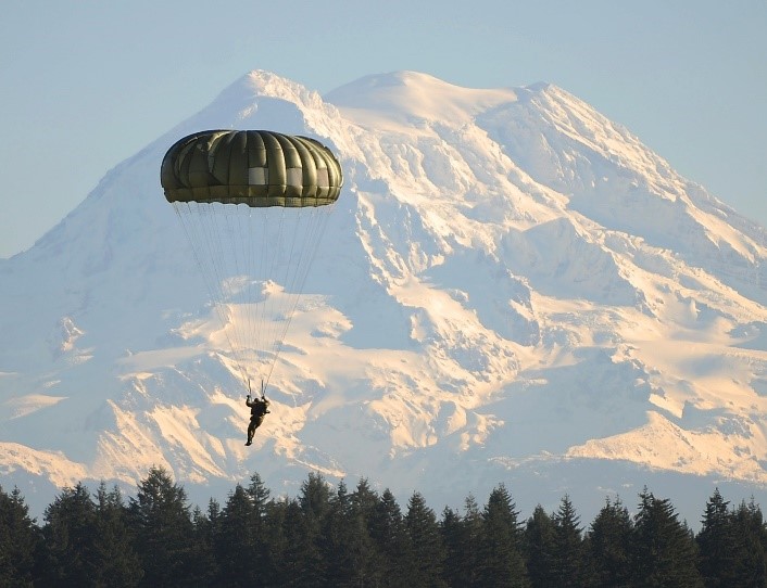 Photographie d'un parachutiste militaire voile déployée. Il se situe dans un paysage montagneux, près de la cime des arbres.